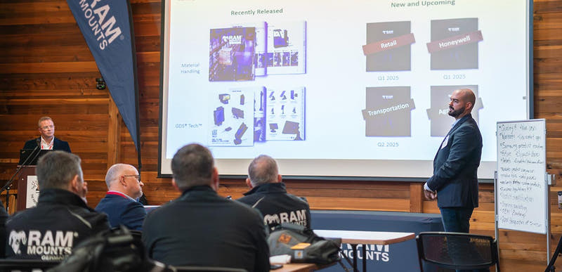 RAM® Mounts team members attending a product presentation and training session at a company event. A presenter stands at the front of the room beside a large projection screen displaying product roadmaps, industry solutions, and upcoming releases, while attendees seated at tables watch the presentation. RAM® Mounts branding is visible throughout the room, and a whiteboard with discussion notes highlights the collaborative, educational nature of the event.