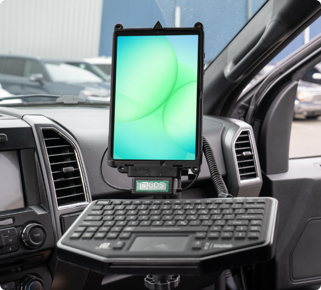 Inside a fleet or patrol vehicle, a Samsung Galaxy Tab A11 is docked in a RAM Mount GDS charging cradle with IntelliSkin, paired with a mounted keyboard tray below. This mobile workstation setup enables full tablet and data entry functionality for dispatch, reporting, or field operations.