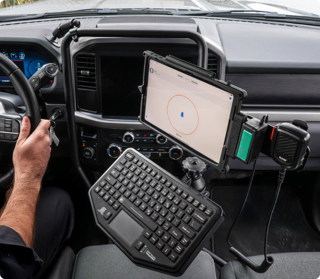 Tablet and mic mounted on a RAM® Tough-Bar system, displaying a push to talk screen with a keyboard below. A person is in the driver’s seat holding onto the wheel.