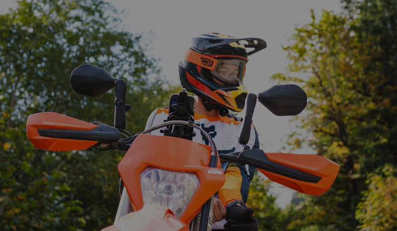 Off-road motorcyclist riding an orange dual-sport motorcycle on a forest trail. The rider wears a full-face helmet, goggles, and protective gear, while a device mount is visible on the motorcycle's handlebars. Trees and natural scenery surround the trail, illustrating adventure riding, powersports navigation, device mounting, and outdoor recreation applications.