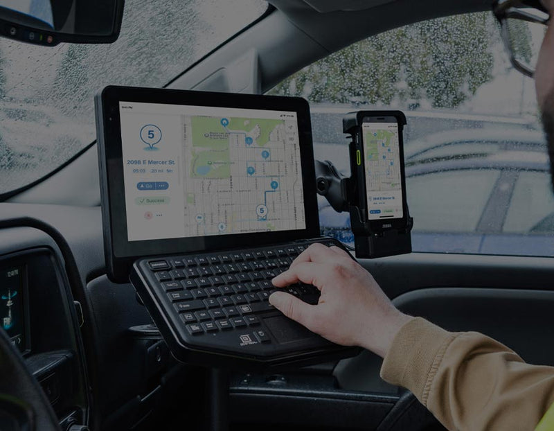 Field service vehicle workstation featuring a rugged tablet and handheld mobile computer mounted inside a truck cab. The tablet displays mapping and route management software and is paired with a compact keyboard for data entry, while a handheld device is docked alongside for quick access. A worker operates the mobile workstation from the driver's seat, illustrating fleet management, route optimization, field service operations, and connected workforce productivity in a commercial vehicle environment.