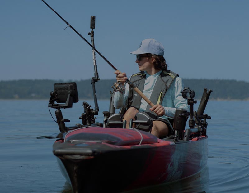 Kayak angler fishing on calm open water with marine electronics mounted throughout the vessel. The kayaker holds a fishing rod while seated in a fully equipped fishing kayak featuring mounted displays, rod holders, and accessory mounts. The scene highlights recreational fishing, marine navigation, hands-free equipment organization, and on-water adventure applications.