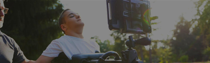 Individual using a wheelchair-mounted tablet system outdoors. The rugged tablet is positioned on an adjustable RAM® mounting arm, providing accessible screen viewing and device control. A caregiver stands nearby while the user interacts with the setup, demonstrating assistive technology solutions that support communication, independence, environmental control, and accessibility for individuals with mobility challenges.