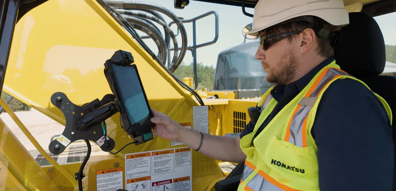 Construction equipment operator using a rugged tablet mounted inside the cab of a heavy machine. The worker, wearing a hard hat, safety glasses, and a high-visibility vest, interacts with a tablet secured to a RAM® mounting system attached to the vehicle frame. The industrial equipment's controls, safety labels, and structural components are visible throughout the cab, illustrating machine guidance, fleet management, jobsite productivity, and digital workflow applications in construction, mining, and heavy 