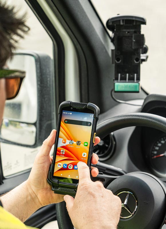 Driver using a rugged Zebra TC73 TC78 handheld mobile computer inside a commercial vehicle. The device, protected by an IntelliSkin® case, is held in front of the steering wheel while an empty GDS® docking station is mounted near the windshield. The screen displays business and productivity applications, illustrating mobile workflow management, fleet operations, field service, logistics, and transportation use cases. The vehicle interior and side mirror are visible in the background.
