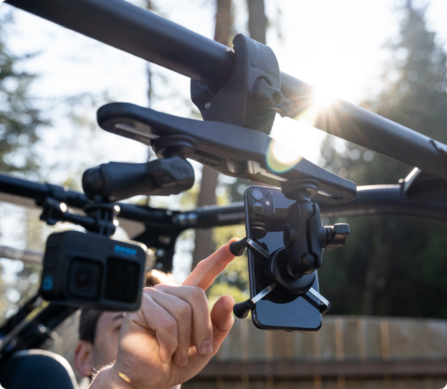 Multiple action cameras and smartphone mounted on RAM Mount multi-device system attached to vehicle roll cage, with driver's hand adjusting mounted smartphone holder while GoPro cameras capture different angles simultaneously.
