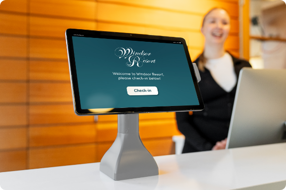 Tablet displaying Windsor Resort check-in welcome screen secured in RAM® Mount countertop kiosk stand at hotel reception desk, enabling self-service guest check-in with hotel staff member visible in background providing customer service support.