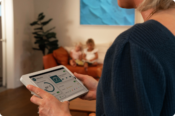 Guest holding tablet with smart home control interface displaying energy monitoring dashboard in modern hotel room, demonstrating RAM® Mount solutions for in-room technology that allows guests to manage lighting, temperature, and entertainment systems with family relaxing in background.