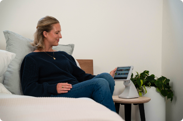 Guest relaxing on hotel bed while viewing tablet secured in RAM® Mount desktop stand on bedside table, displaying room control interface for managing lighting, temperature, and entertainment in modern hospitality environment with minimalist decor.