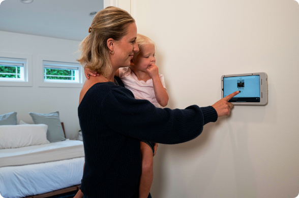 Mother holding child while interacting with tablet displaying smart home controls secured in RAM® Mount wall-mounted holder in hotel room, demonstrating intuitive touch interface for adjusting room settings and amenities in family-friendly hospitality environment.