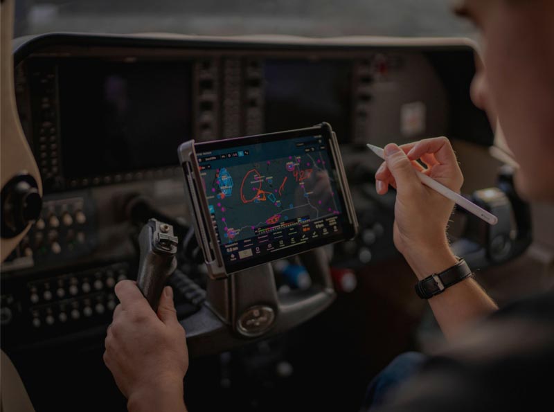 Pilot using a tablet mounted to an aircraft control yoke inside a modern cockpit. The tablet displays aviation navigation charts and flight information while the pilot references the screen using a stylus. Flight instruments, avionics displays, and cockpit controls surround the workstation, illustrating electronic flight bag (EFB) applications for navigation, flight planning, situational awareness, and aviation workflow management.