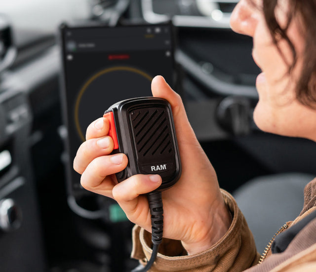Law enforcement officer using a RAM® Remote Speaker Mic with USB-C beside an Apple Device mounted inside a patrol vehicle. The tablet is wrapped in an IntelliSkin® Next Gen protective sleeve and docked in a RAM® GDS® Uni-Conn™ Dock mounted to a RAM® No-Drill™ Vehicle Base, creating a rugged push-to-talk tablet solution.