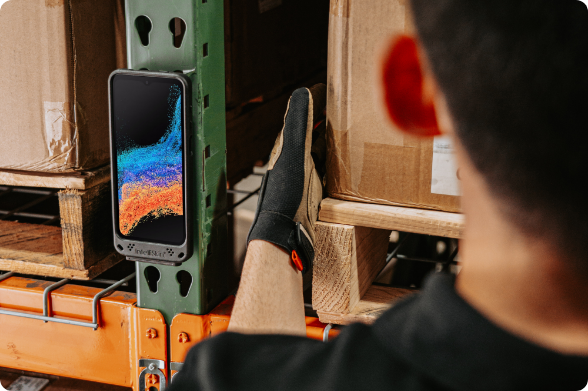 Rugged smartphone with protective case secured in RAM® magnetic holder mounted on industrial equipment frame in warehouse setting, enabling hands-free device access during logistics operations with worker visible in background performing tasks.