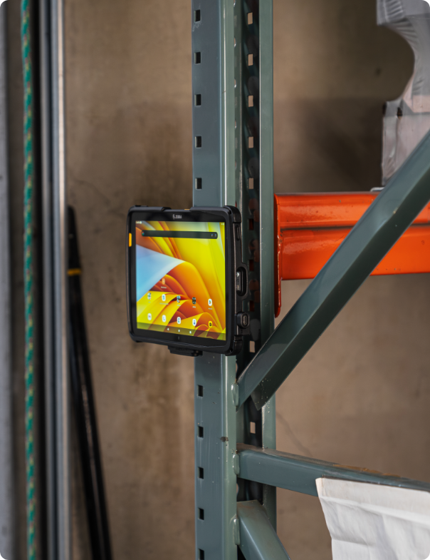 Zebra rugged tablet displaying colorful interface secured in RAM® Magnetic Mount holder attached to warehouse storage rack vertical post, demonstrating hands-free access to inventory management applications during logistics operations in industrial facility with storage shelves visible in background.