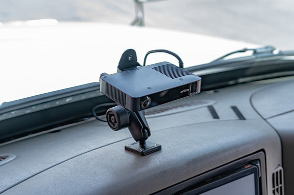 Motive AIDC+ camera mounted to a RAM® dashboard mounting system inside a commercial vehicle. The camera is positioned above the dashboard with the driver’s hand visible on the steering wheel, while the road and surrounding landscape appear motion-blurred through the windshield, highlighting fleet safety and mobile device mounting solutions.