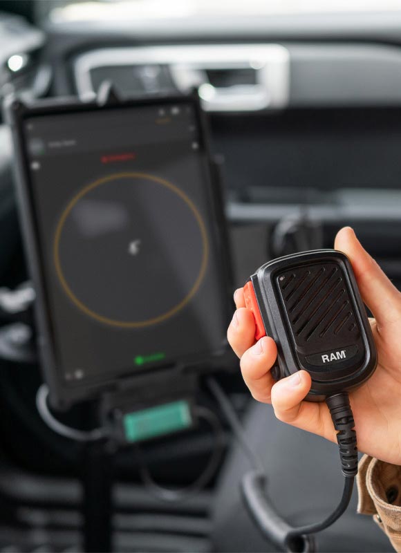 Hand holding a RAM® push-to-talk microphone inside a vehicle, with a mounted tablet visible in the background. The microphone features a rugged, ergonomic design and is connected by a coiled cable, supporting clear voice communication for fleet, public safety, transportation, and field service operations. The tablet is docked on the vehicle dashboard, highlighting an integrated mobile workstation setup for connected communications and workflow management.