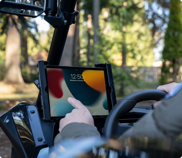 Tablet secured in RAM Mount holder attached to off-road vehicle roll bar, displaying colorful lock screen showing 12:50 time while driver's hand interacts with the mounted device during outdoor expedition.