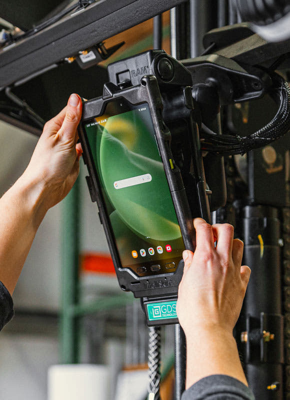 Worker docking a rugged tablet into a RAM® GDS® vehicle or industrial mounting system. The tablet, protected by an IntelliSkin® enclosure, is being secured into a powered dock mounted to heavy-duty equipment. Hands guide the device into place while integrated charging and connectivity components support reliable tablet operation in warehouse, manufacturing, fleet, and industrial environments. Background machinery is softly blurred to emphasize the docking process.