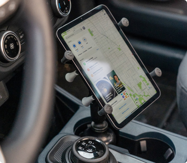 Tablet displaying detailed street map with navigation interface secured in RAM® X-Grip® holder mounted in vehicle cup holder area, positioned for optimal viewing angle during GPS navigation and route planning.