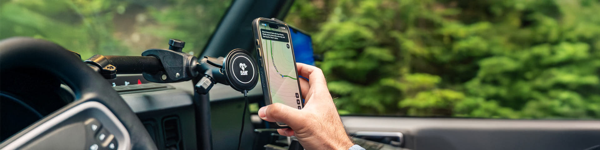 Driver interacting with a smartphone mounted inside a vehicle using a RAM® magnetic wireless charging mount. The phone displays a navigation map while mounted near the dashboard, and a hand is shown removing or repositioning the device. The vehicle interior is visible in the foreground, with a blurred green outdoor landscape through the window, illustrating convenient in-vehicle navigation and hands-free device access.