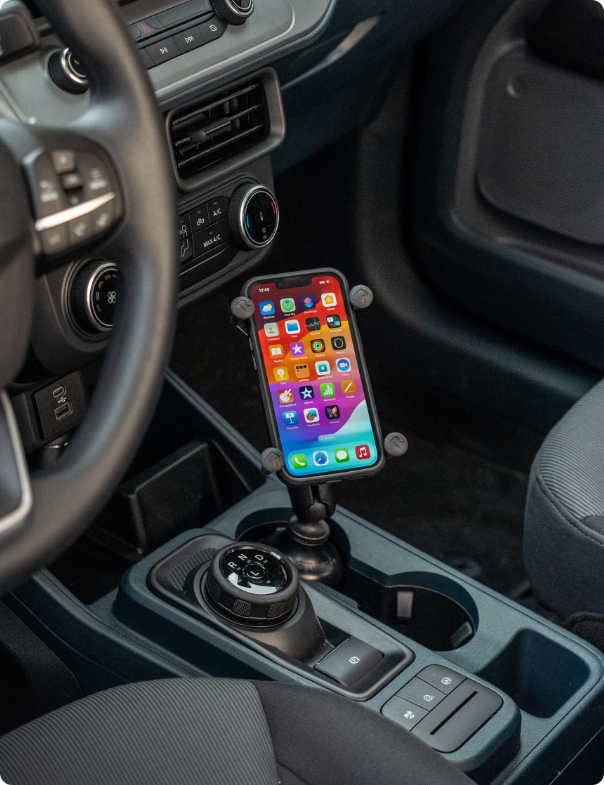 iPhone showing colorful home screen with app icons secured in RAM® X-Grip® holder mounted in vehicle cup holder between center console controls, providing easy access to smartphone functions while maintaining driver safety.